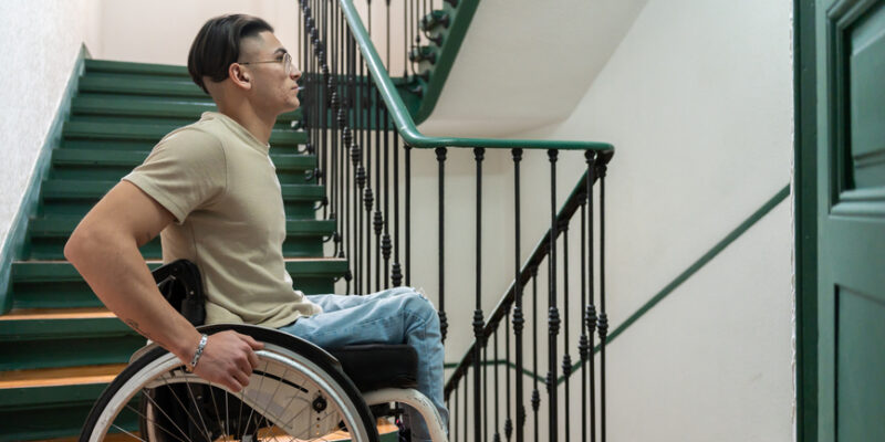 Portrait Looking At The Camera Of A Young Man With A Disability Who On The Stairs Of A Building. Wheelchair User In The Corridor Of A Building Without Accessibility For People With Disabilities.